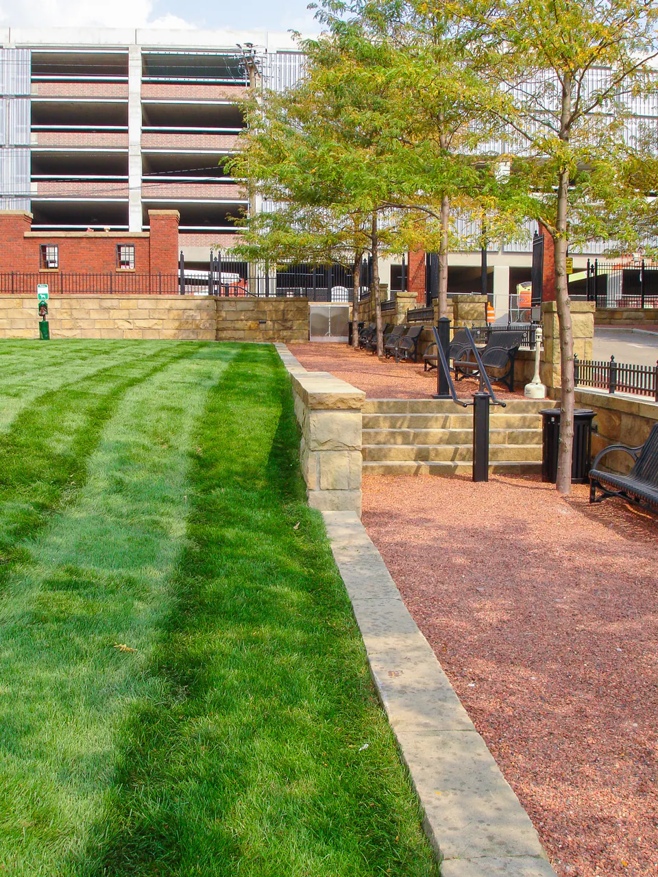 Narrow courtyard view with lawn, brick walk, stone wall, railings, trees, and parking structure beyond.